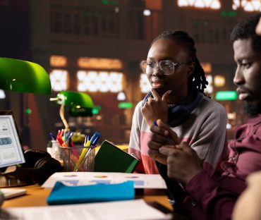 A Black woman with glasses and a Black man reviewing user data charts on a screen and paper documents in a dimly lit studio.