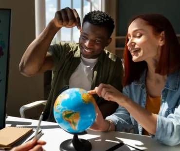 A smiling Black man and a redhead woman sitting at a desk together, pointing at a small desk globe while discussing a project.