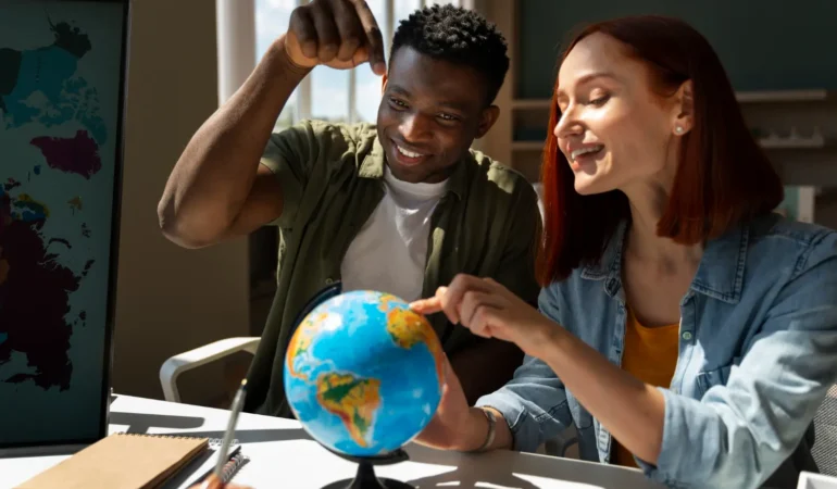 A smiling Black man and a redhead woman sitting at a desk together, pointing at a small desk globe while discussing a project.