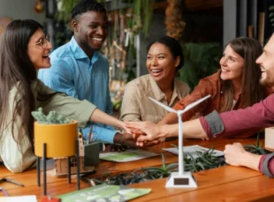 A diverse group of five smiling colleagues stacking their hands together over a wooden table featuring a small model wind turbine and green plants.