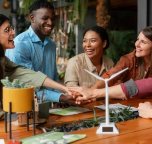 A diverse group of five smiling colleagues stacking their hands together over a wooden table featuring a small model wind turbine and green plants.