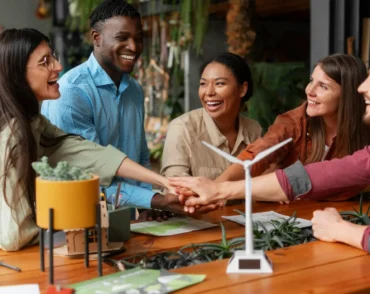A diverse group of five smiling colleagues stacking their hands together over a wooden table featuring a small model wind turbine and green plants.