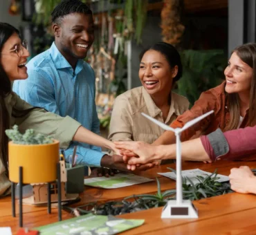 A diverse group of five smiling colleagues stacking their hands together over a wooden table featuring a small model wind turbine and green plants.
