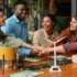 A diverse group of five smiling colleagues stacking their hands together over a wooden table featuring a small model wind turbine and green plants.