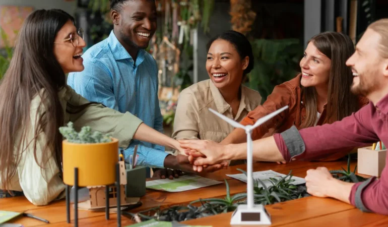 A diverse group of five smiling colleagues stacking their hands together over a wooden table featuring a small model wind turbine and green plants.