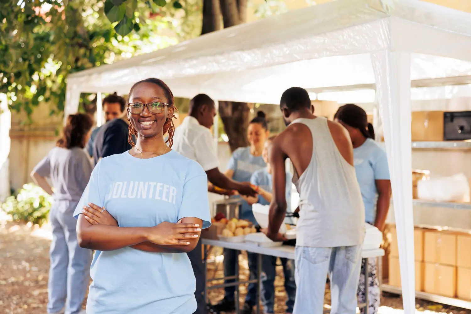 Community volunteers working at an outdoor donation drive A smiling young woman with glasses wearing a blue "VOLUNTEER" t-shirt, standing with arms crossed in front of an outdoor community service event.
